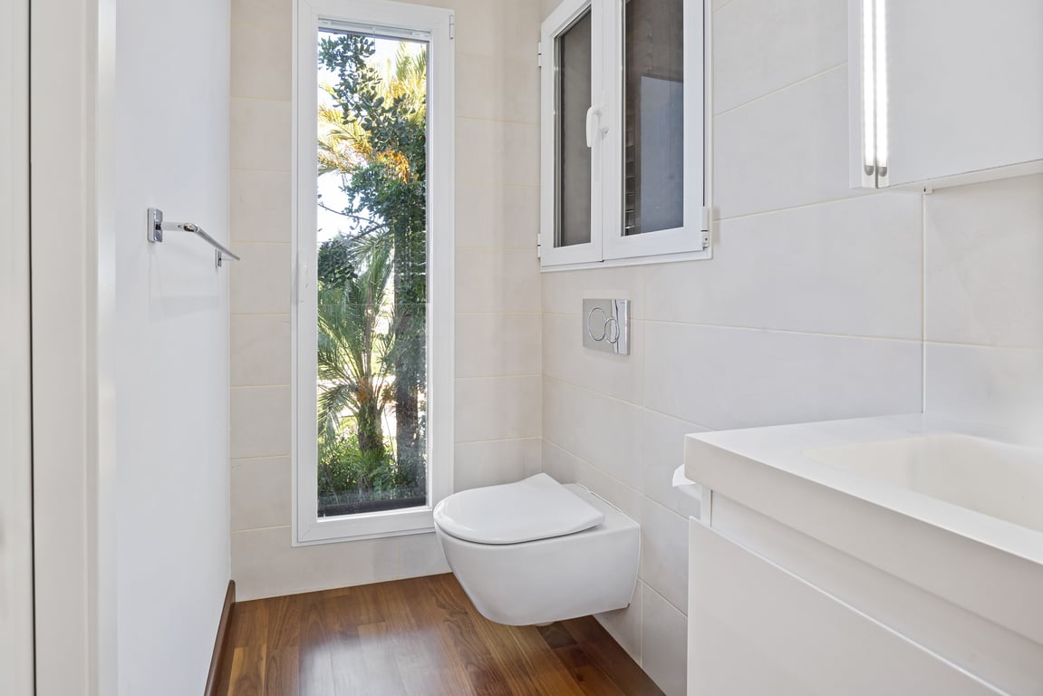 Modernisiertes Badezimmer mit wandhängendem WC, Holzboden und einem vertikalen Fenster mit Blick auf den mediterranen Garten.