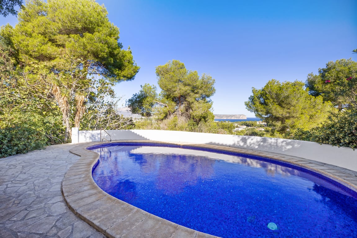 Nierenförmiger Privatpool mit blauem Mosaik, umgeben von einer Natursteinterrasse und Pinien mit weitem Blick auf das Mittelmeer.