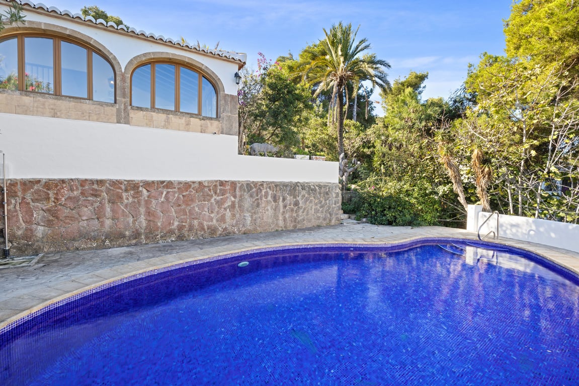 Privater Pool mit blauen Mosaikfliesen, eingefasst von einer rustikalen Natursteinmauer und der verglasten Naya der Villa inmitten mediterraner Bepflanzung.