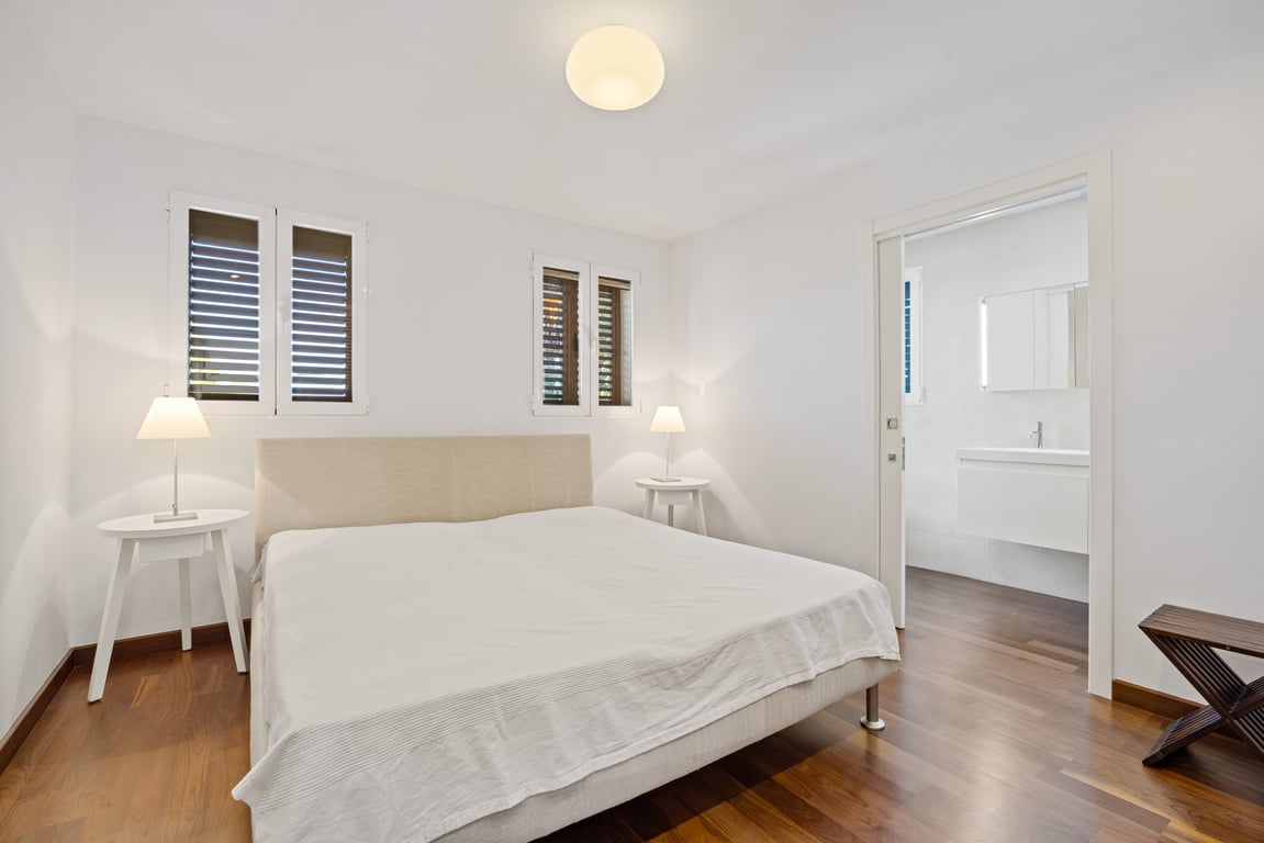 Renoviertes Schlafzimmer mit dunklem Holzboden, Stoffkopfteil und Schiebetür zu einem modernen En-Suite-Badezimmer mit weißem Waschtisch.