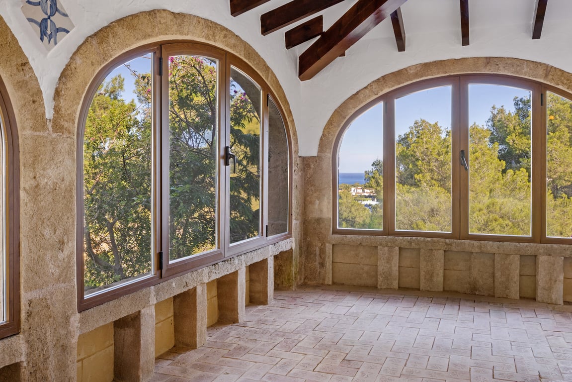 Verglaste Naya mit Tosca-Steinbögen, sichtbaren Holzbalken und handgefertigten Tonfliesen. Die Fenster bieten einen Blick auf das Meer und die grüne Landschaft.