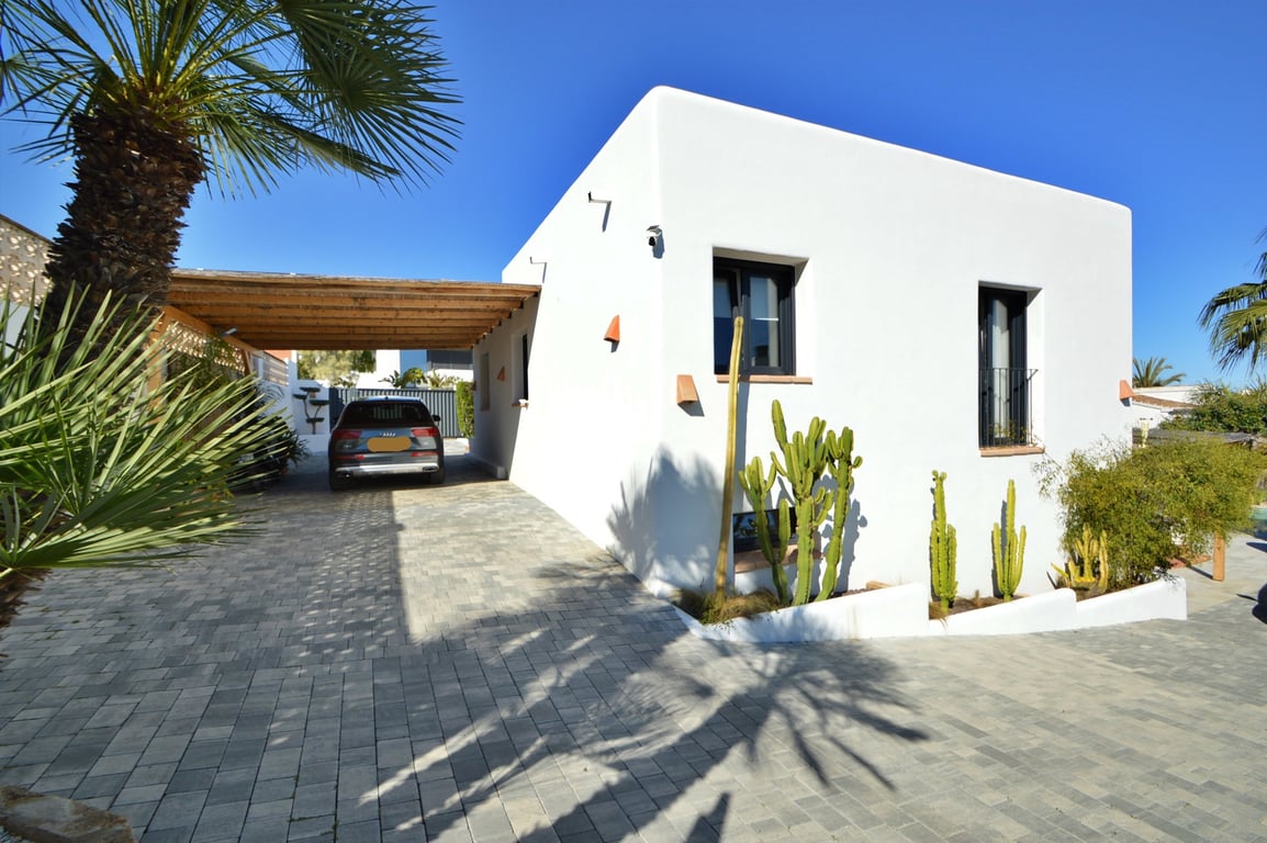 Außenansicht der Villa im Ibiza-Stil mit Holz-Carport, gepflasterter Einfahrt und mediterraner Bepflanzung mit Kakteen und Palmen.