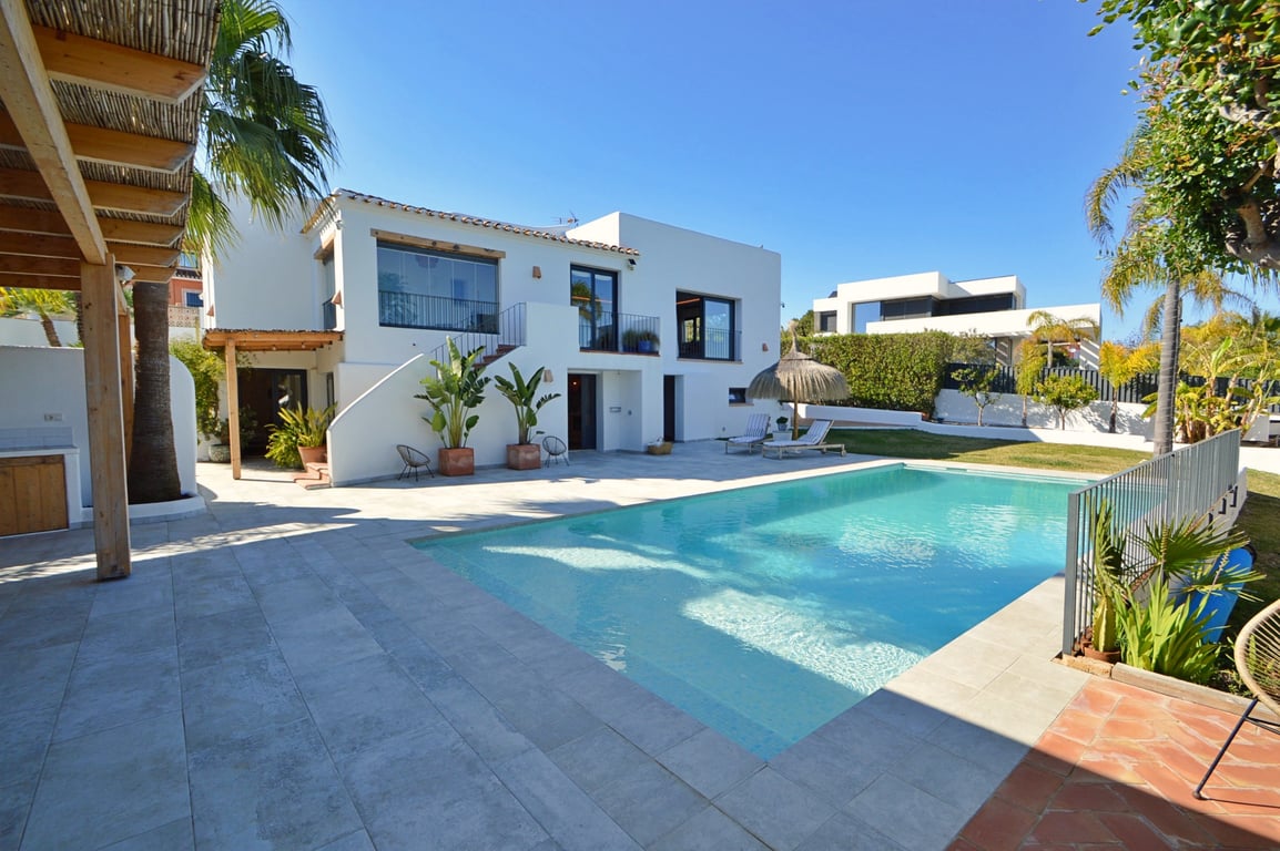 Außenansicht der Villa mit großem Pool, heller Steinterrasse, Holzpergola und weißer Fassade im Ibiza-Stil mit verglaster Naya im Obergeschoss.