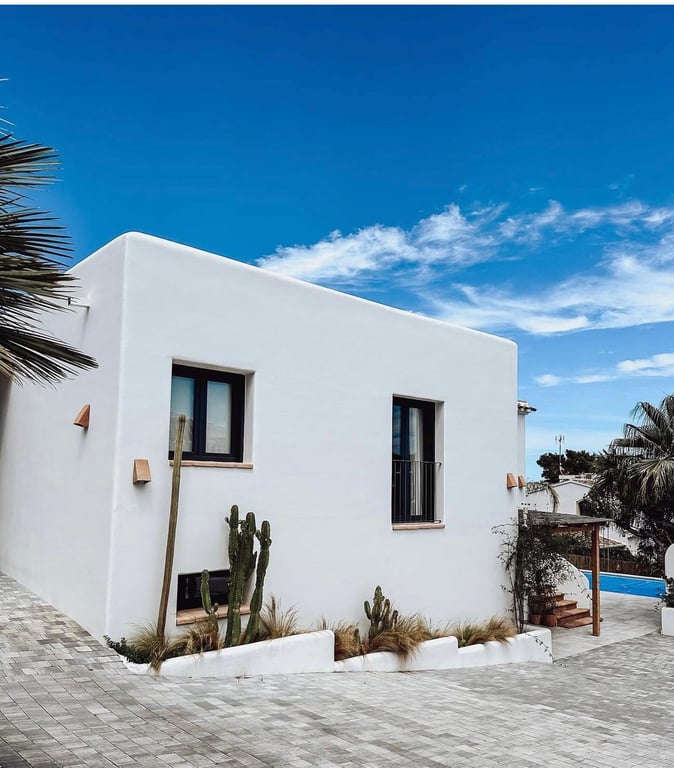 Außenansicht im ibizenkischen Stil mit glatten weißen Wänden, dunklen Fensterrahmen und einem Kakteengarten neben der gepflasterten Zufahrt.