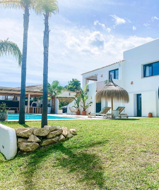 Außenbereich im Ibiza-Stil mit Rasenfläche, Pool und Reet-Sonnenschirm. Im Hintergrund ist die Außenküche mit traditionellem Pizzaofen erkennbar.
