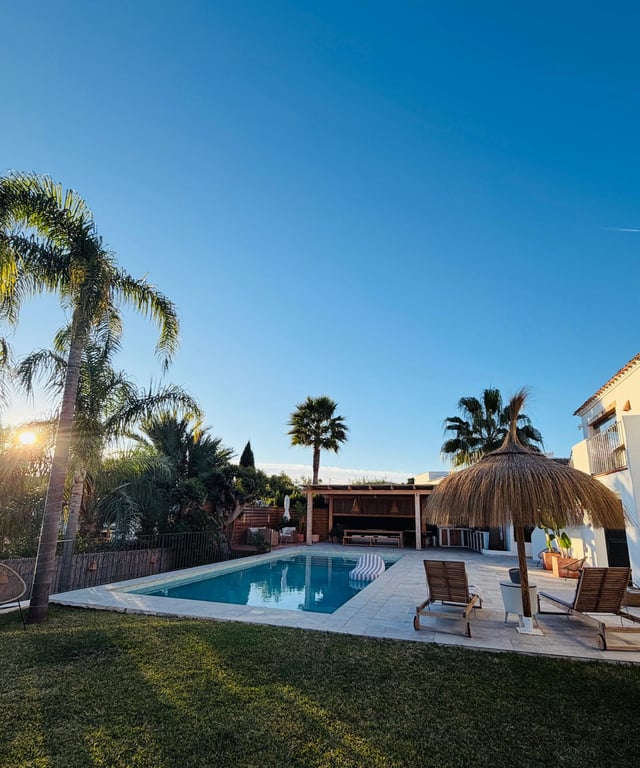 Außenbereich mit Pool, Steinterrasse und Sonnenschirm aus Reet. Im Hintergrund befinden sich eine überdachte Außenküche und ein Essbereich inmitten von gepflegten Palmen.