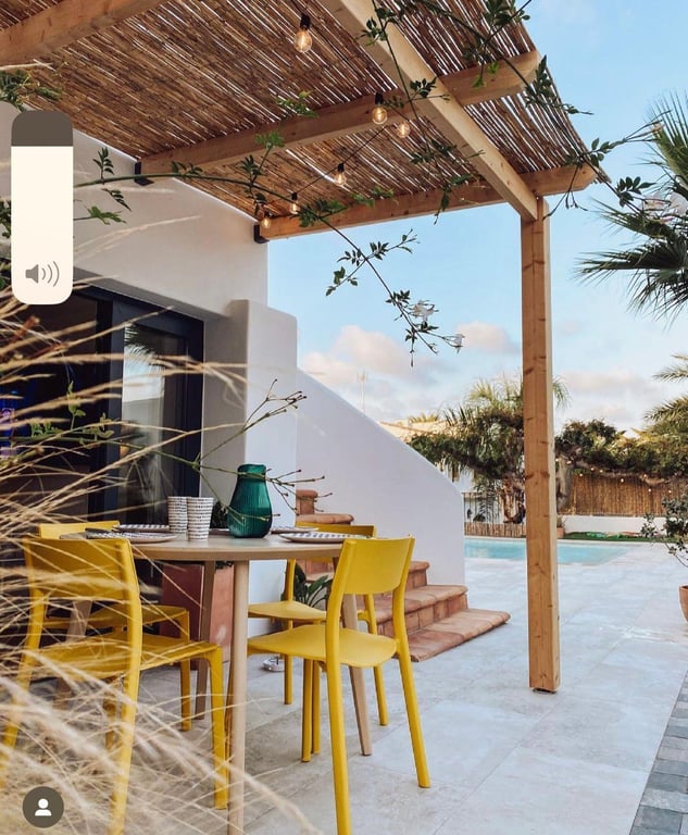 Außenessbereich unter einer Holzpergola mit Schilfdach und Lichterketten. Gelbe Designstühle auf einer Steinterrasse mit Blick auf den Pool und Garten.