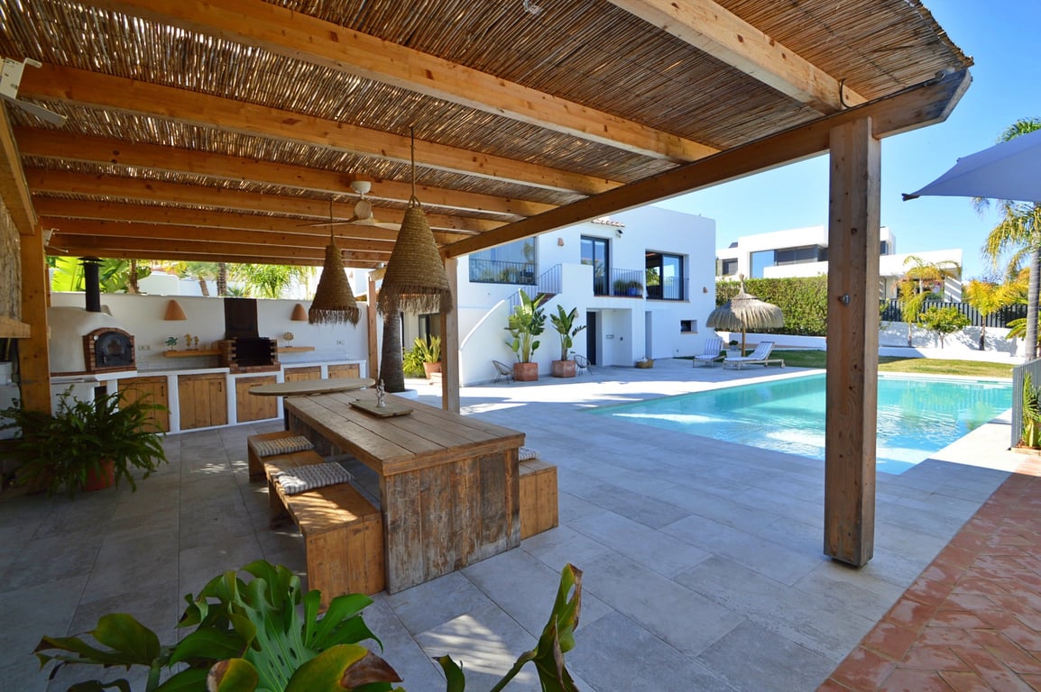 Außenküche unter einer Holzpergola mit Pizzaofen, rustikalem Essbereich und Blick auf den Salzwasserpool und die weiße Villa im Ibiza-Stil.