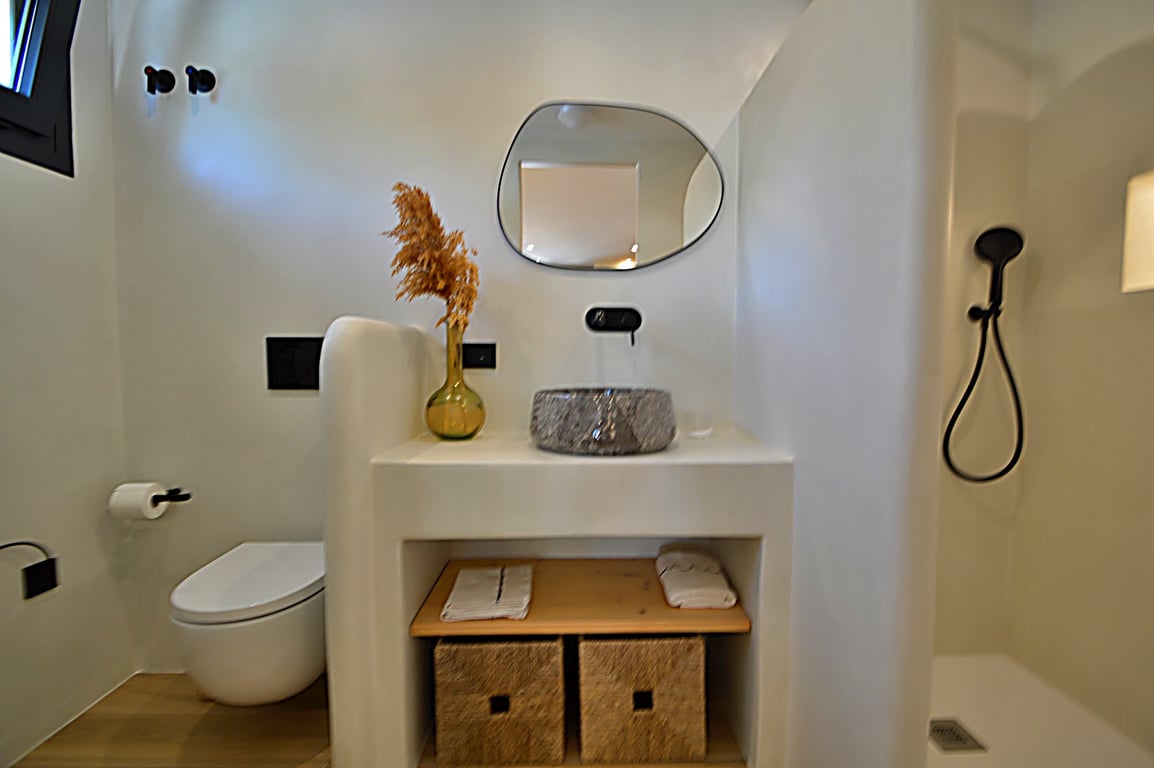 Badezimmer im Ibiza-Stil mit Mikrozementwänden, Steinwaschbecken und mattschwarzen Armaturen. Ein organischer Spiegel und eingebaute Regale mit Weidenkörben sorgen für modernes Flair.
