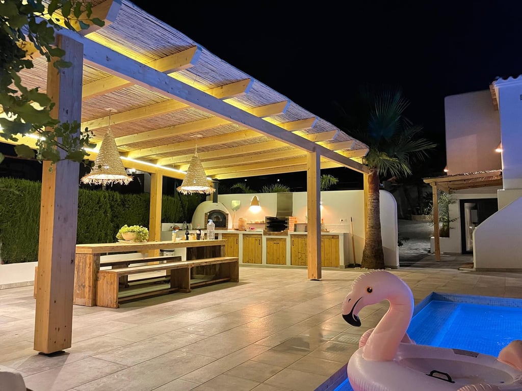 Beleuchtete Außenküche mit Holzofen, Grill und Essbereich unter einer Holzpergola mit Schilfdach neben dem Poolbereich.