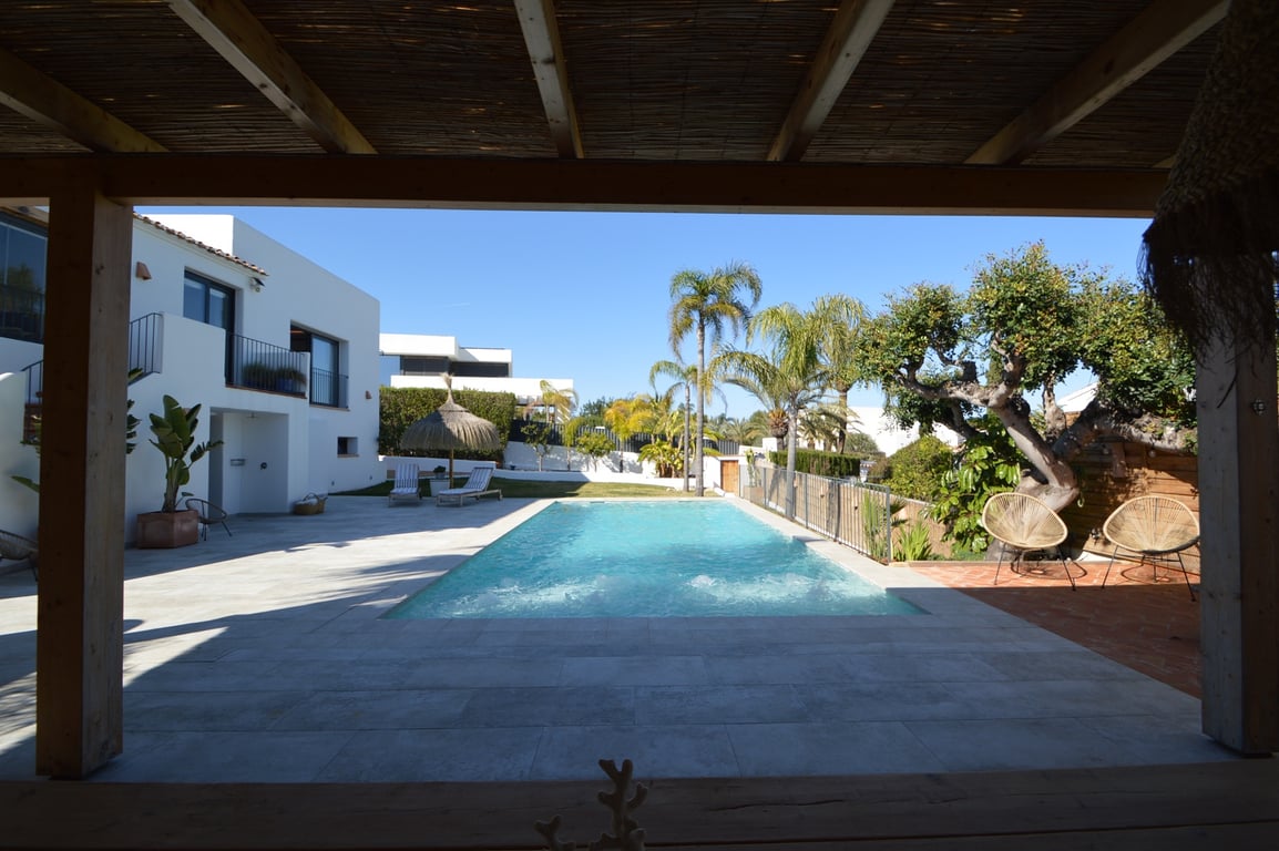 Blick von der Holzpergola auf den Salzwasserpool, umgeben von hellen Natursteinterrassen, Palmen und der weißen Villa im ibizenkischen Stil.
