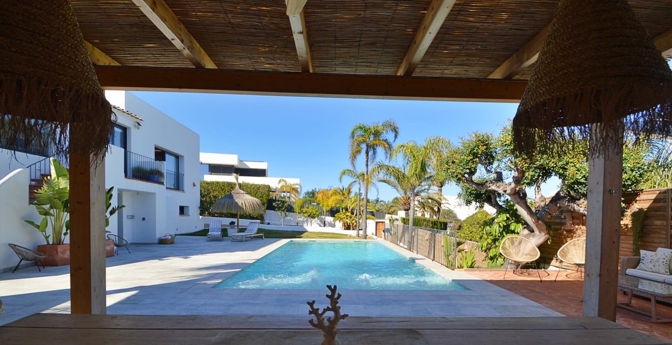 Blick von der schattigen Terrasse mit Schilfrohrdach auf den Salzwasserpool und den mediterranen Garten mit Palmen.