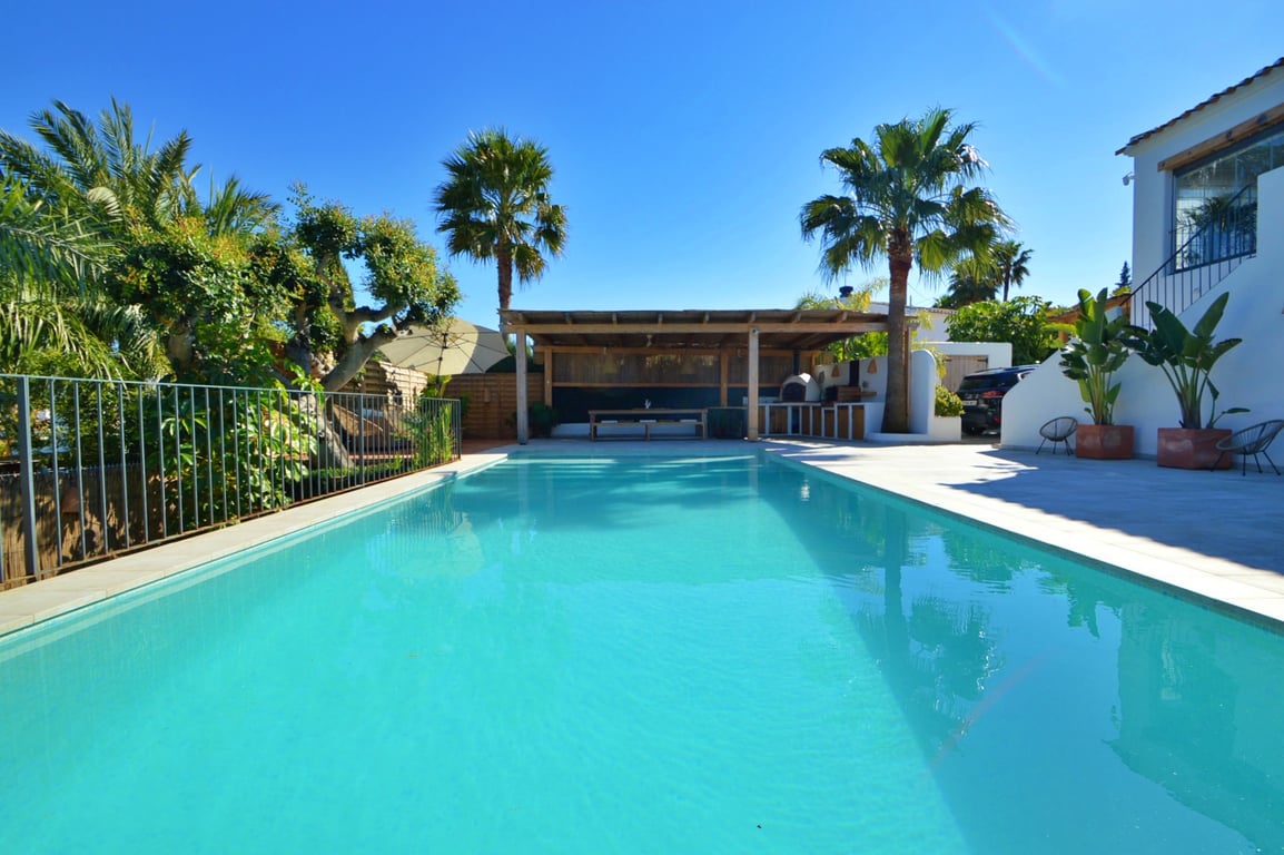 Großer rechteckiger Pool mit weitläufiger Sonnenterrasse, einem überdachten Essbereich unter einer Holzpergola und mediterraner Bepflanzung mit Palmen.