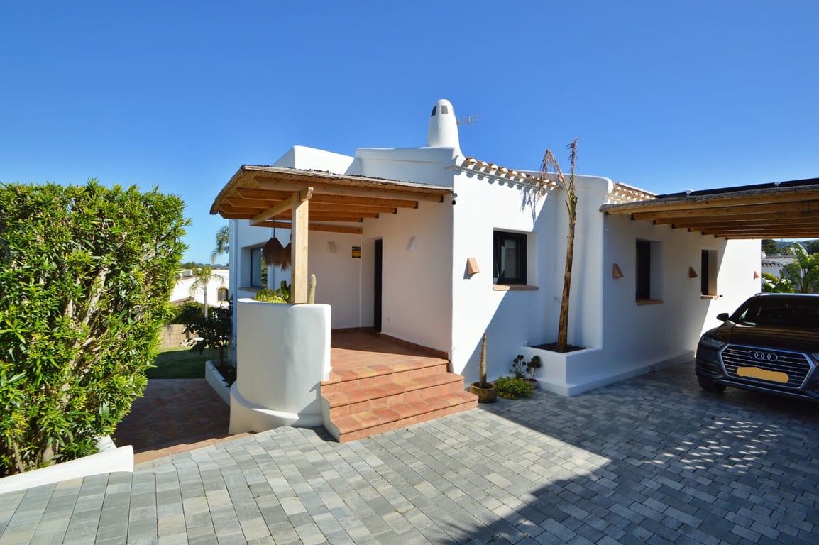 Haupteingang im ibizenkischen Stil mit Holzpergola und Schilfdach, Terrakotta-Stufen und gepflasterter Einfahrt mit seitlichem Carport.