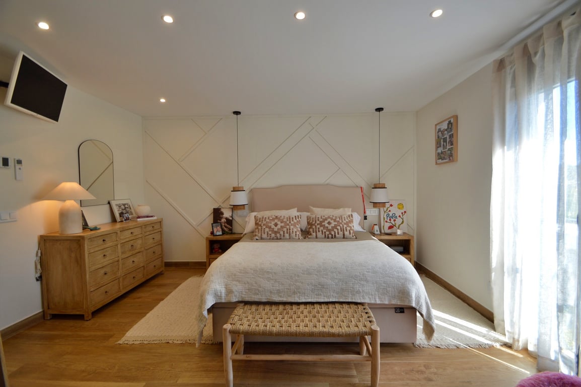 Hauptschlafzimmer mit geometrischer Akzentwand, hellem Holzfußboden und Deckenspots. Zur Ausstattung gehören eine Holzkommode, Hängeleuchten und eine geflochtene Sitzbank.