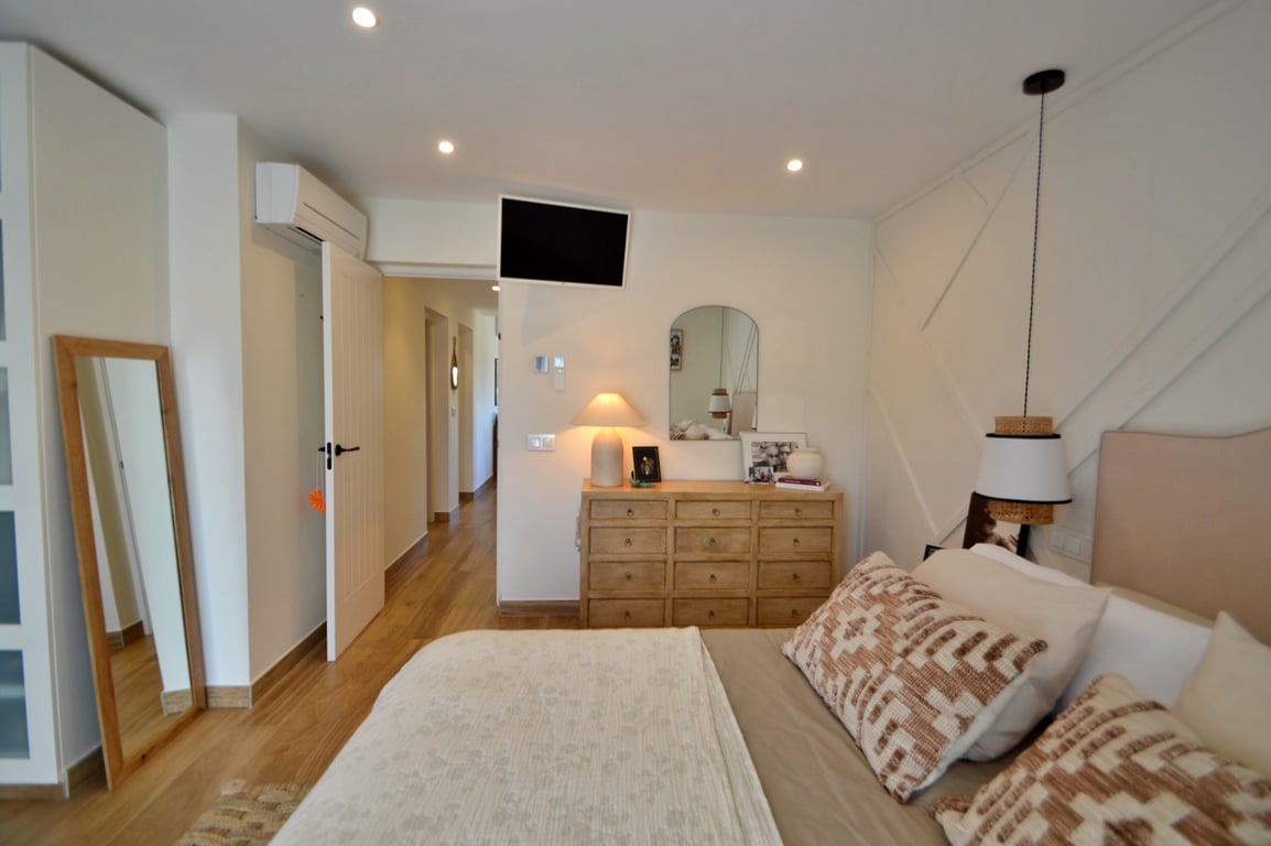 Helles Schlafzimmer mit Bodenbelag in Holzoptik, geometrischer Wandgestaltung, Klimaanlage und Naturholzmöbeln wie einer Kommode und einem Standspiegel.