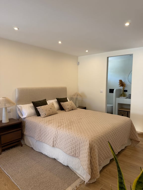 Helles Schlafzimmer mit Doppelbett, gepolstertem Kopfteil und Nachttischen aus Holz. Integriertes En-Suite-Badezimmer mit modernen Armaturen und Einbaustrahlern.