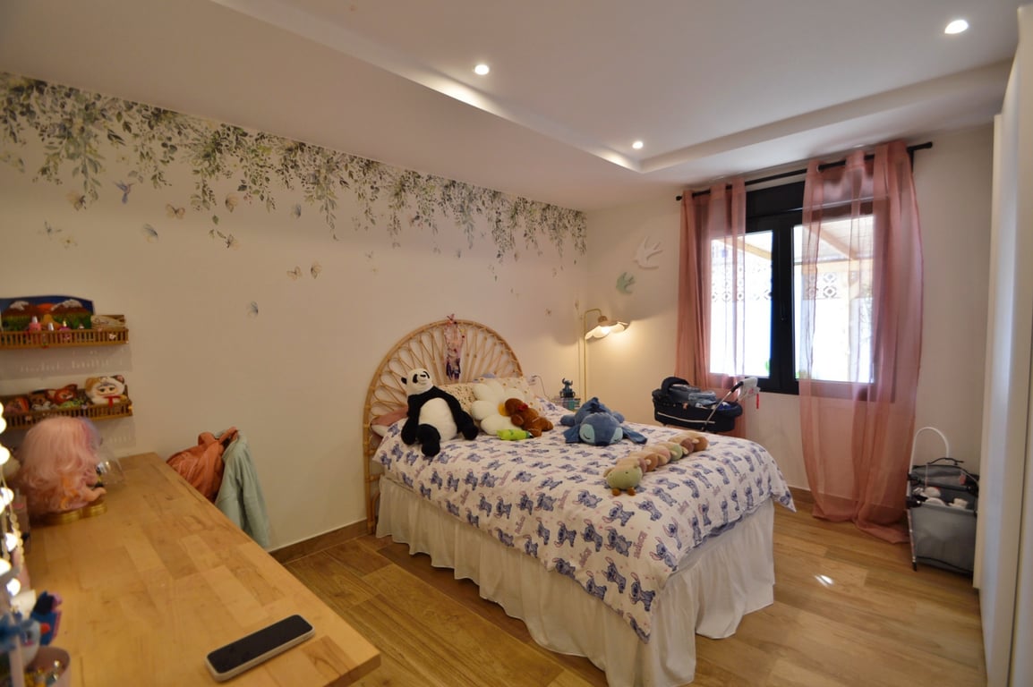 Helles Schlafzimmer mit Rattan-Kopfteil, botanischer Wandgestaltung und Holzboden. Einbaustrahler und natürliche Lichtquellen sorgen für ein einladendes Ambiente.