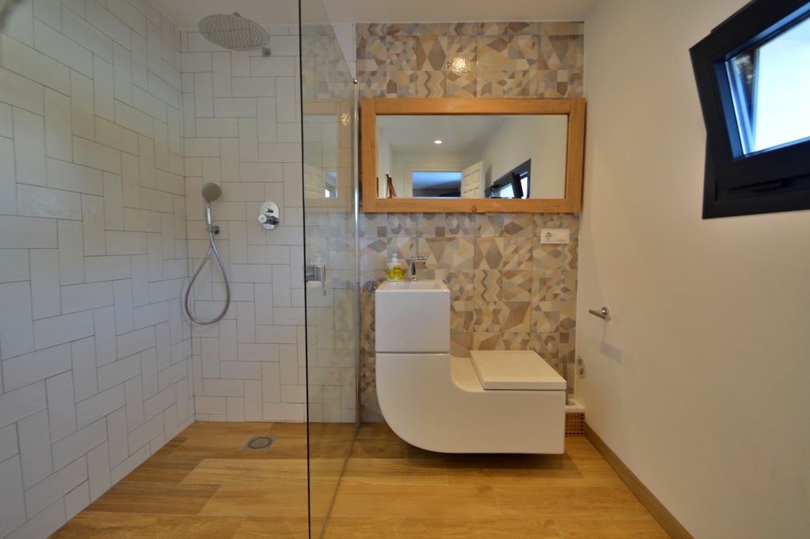 Modernes Badezimmer mit ebenerdiger Dusche, Glaswand und einer kombinierten Waschbecken-WC-Einheit. Akzentuiert durch geometrische Fliesen und einen Spiegel mit Holzrahmen.