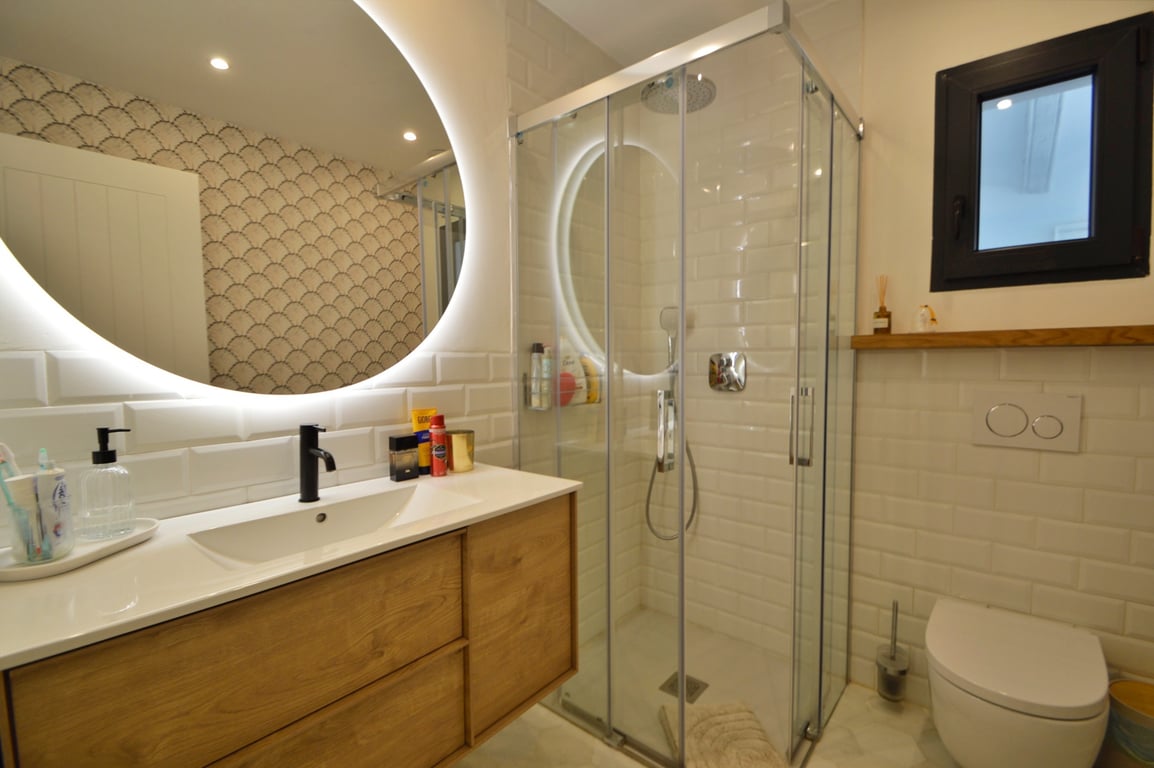 Modernes Badezimmer mit hinterleuchtetem Rundspiegel, Holzwaschtisch und Eckdusche mit klassischen weißen Metrofliesen.