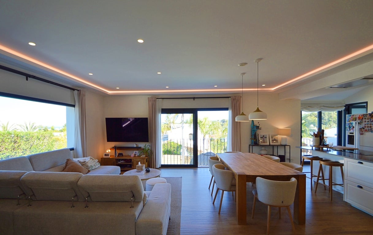 Offener Wohn- und Essbereich mit moderner LED-Deckenbeleuchtung, hochwertigem Holzboden und großen Fensterfronten, die viel natürliches Licht hereinlassen.