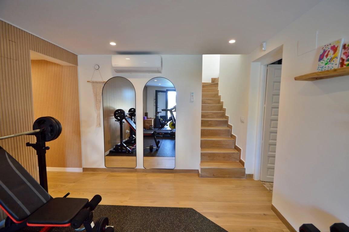 Privater Fitnessraum mit Rundbogenspiegeln, Boden in Holzoptik, Klimaanlage und integrierter Treppe, die funktionales Design mit Komfort verbindet.
