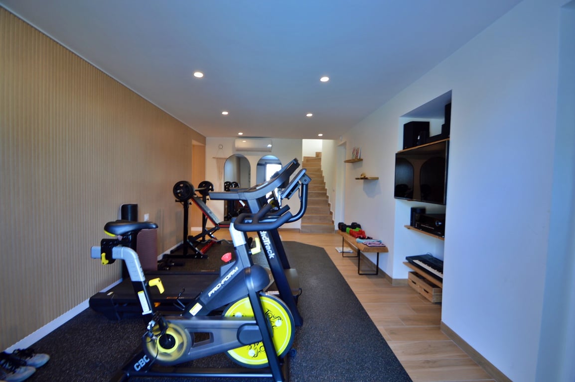 Privater Fitnessraum mit Spinning-Bike, Laufband und Hantelbank, ausgestattet mit Holzpaneelwand, Deckenspots und integrierter Multimedia-Nische.