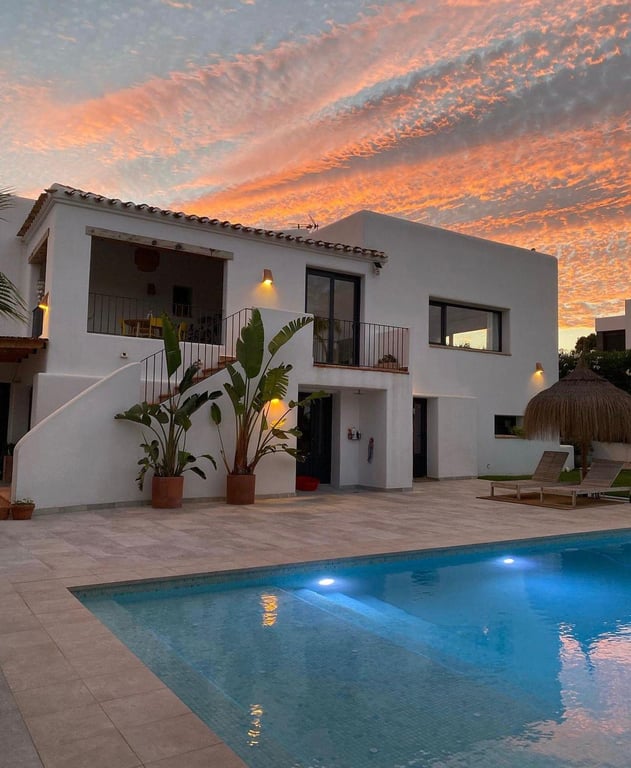 Villa im Ibiza-Stil bei Dämmerung mit beleuchtetem Salzwasserpool, Natursteinterrasse und mediterraner Bepflanzung vor einem spektakulären Abendhimmel.