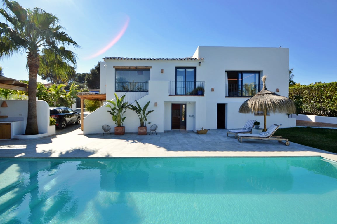 Weiße Fassade im Ibiza-Stil vor einem großen Salzwasserpool, mit Steinterrasse, Palmen und einem Liegebereich mit traditionellem Strohschirm.