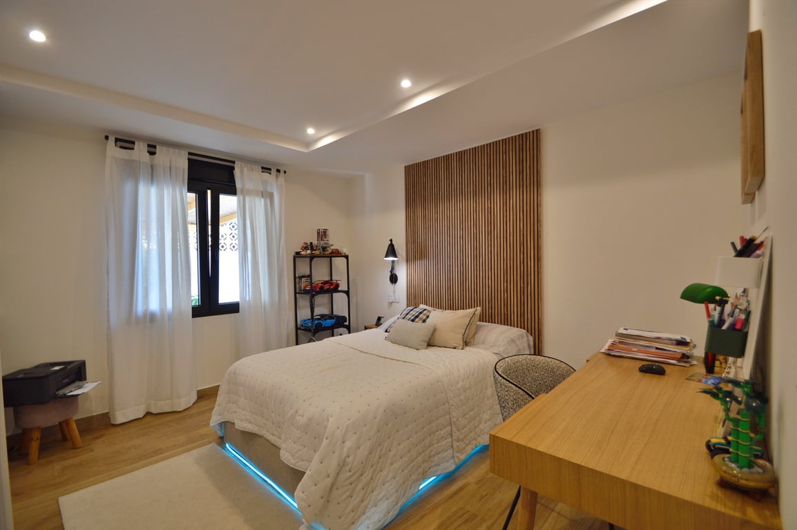 Zeitgemäßes Schlafzimmer mit Holzlamellen-Kopfteil, Schreibtisch und Bett mit blauer LED-Akzentbeleuchtung. Mit Holzboden und Deckeneinbaustrahlern ausgestattet.