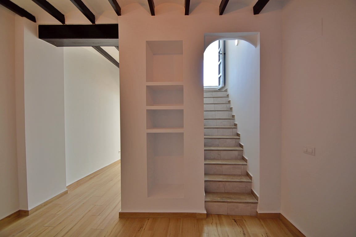 Innenflur mit sichtbaren dunklen Holzbalken, integrierten Wandnischen und einer gefliesten Treppe, die helles Tageslicht in den Raum lässt.