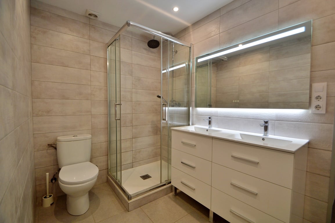 Modernes Badezimmer mit Doppelwaschtisch, weißem Unterschrank, hinterleuchtetem Spiegel und einer Eckduschkabine aus Glas mit hellen Fliesen.