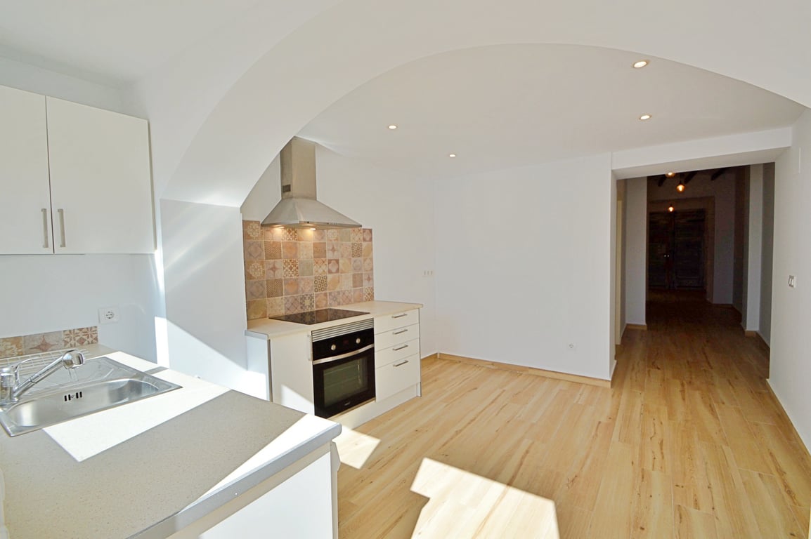 Renovierte Küche mit weißen Schränken, dekorativem Fliesenspiegel und Einbaugeräten. Heller Holzboden, Deckenspots und ein eleganter Rundbogen zum Wohnbereich.