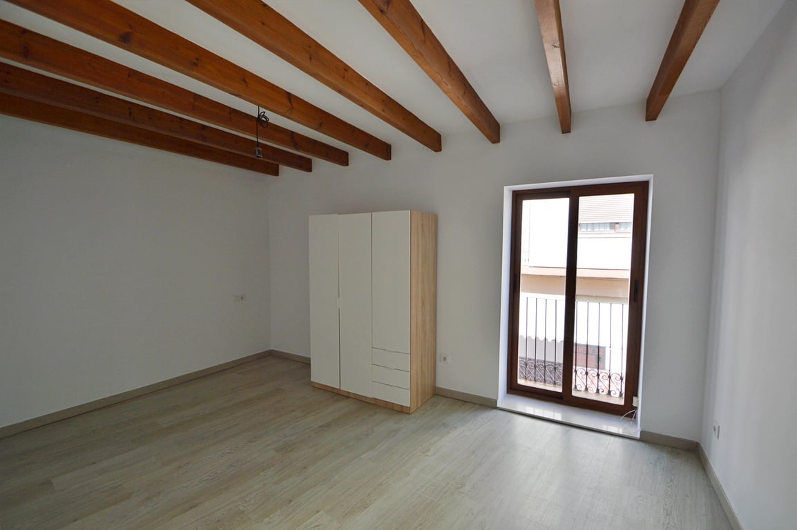Schlafzimmer mit sichtbaren Holzbalken, hellem Laminatboden und modernem Kleiderschrank. Eine Glasschiebetür führt auf einen privaten Balkon mit Eisenreling.