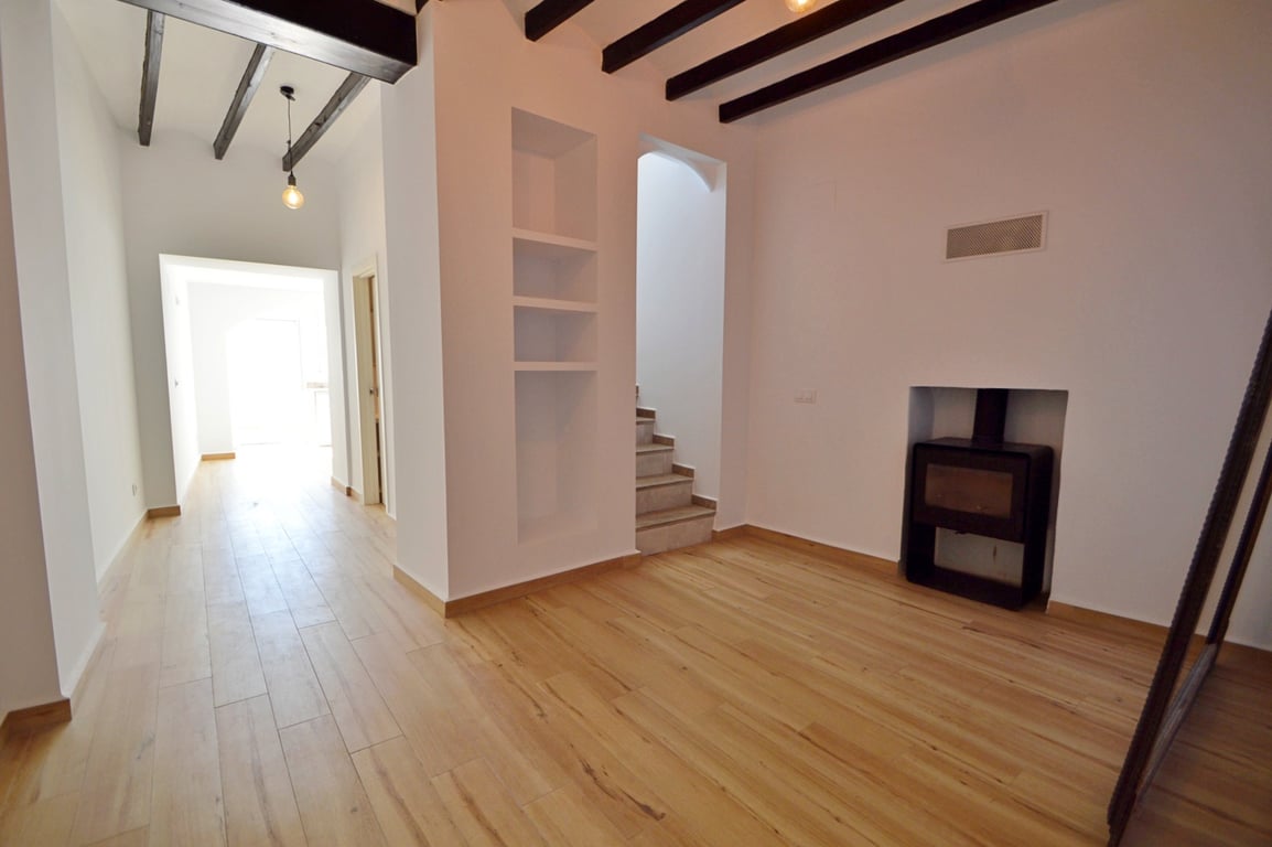 Wohnbereich mit dunklen Deckenbalken, hellem Boden in Holzoptik und integriertem Kaminofen. Einbauregale und eine Treppe ergänzen den offenen Grundriss.