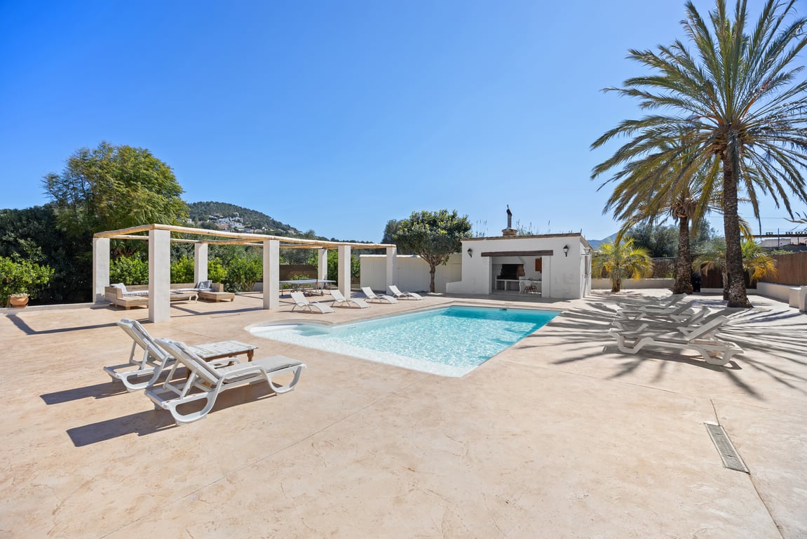 Ein 5x10m großer Pool mit weitläufiger Pflasterterrasse, voll ausgestatteter Außenküche, Holzpergola und mediterranen Palmen unter blauem Himmel.