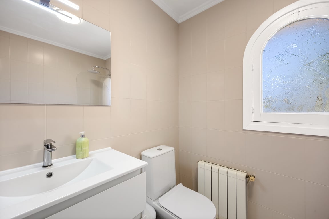 Funktionales Badezimmer mit neutralen Fliesen, weißem Waschtisch, Heizkörper und einem Rundbogenfenster mit Milchglas für natürliches Licht.