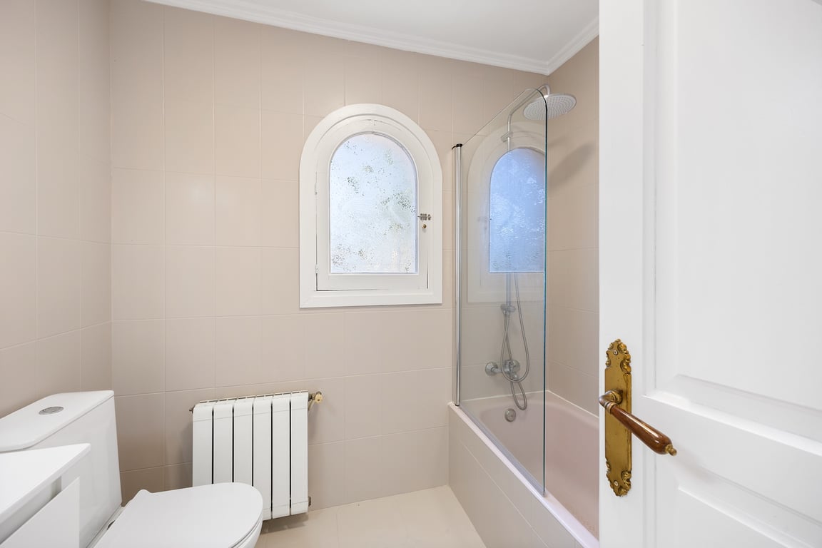 Funktionelles Badezimmer mit Badewanne, Glastrennwand und einem markanten Rundbogenfenster. Ausgestattet mit Heizkörper und Fliesen in neutralen Tönen.
