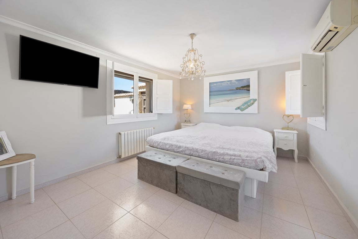 Geräumiges Schlafzimmer mit weißen Fensterläden, Wand-TV und Klimaanlage. Ausgestattet mit einem großen Bett, Polsterhockern und hellem Fliesenboden.