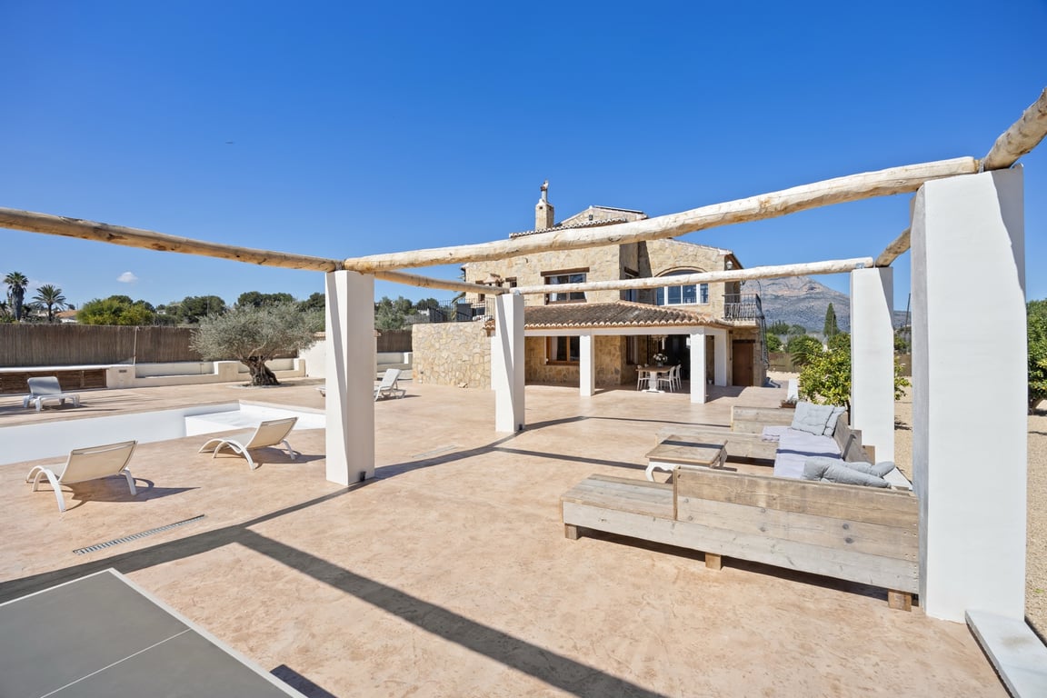 Großzügige Außenterrasse mit Holzbalken-Pergola, Loungebereich und Blick auf die Steinfassade der Villa sowie den Berg Montgó im Hintergrund.
