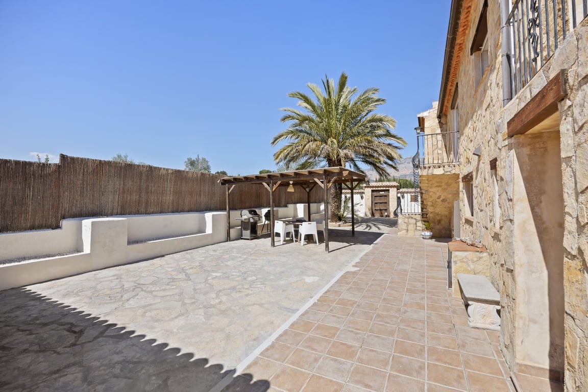 Großzügige Außenterrasse mit Steinpflasterung, einer Holzpergola mit Grillbereich und der charakteristischen Natursteinfassade der Villa.