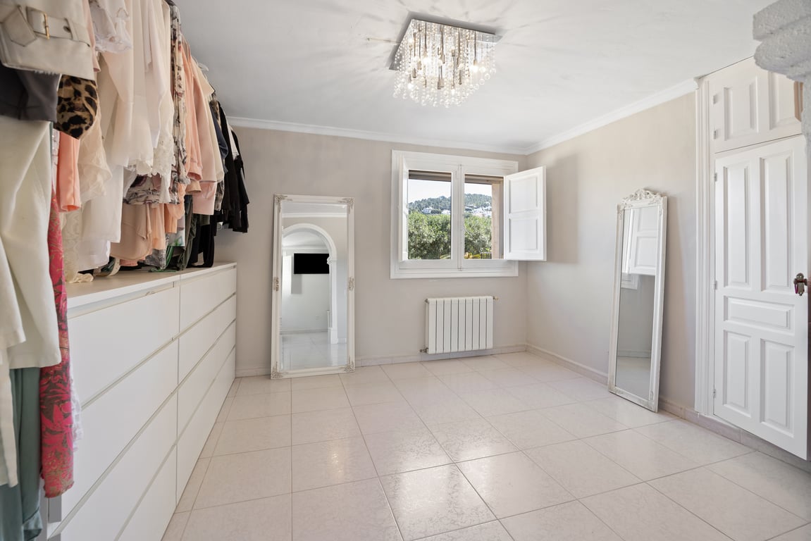Heller begehbarer Kleiderschrank mit weißer Kommode, Ganzkörperspiegeln und Kristallleuchter. Der Raum bietet Zentralheizung und einen Blick ins Grüne durch das Fenster.