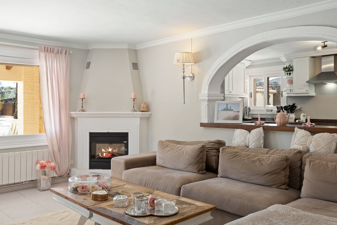 Heller Wohnbereich mit Eckkamin, modularem Sofa in Naturtönen und einem dekorativen Rundbogen mit Blick in die Küche.