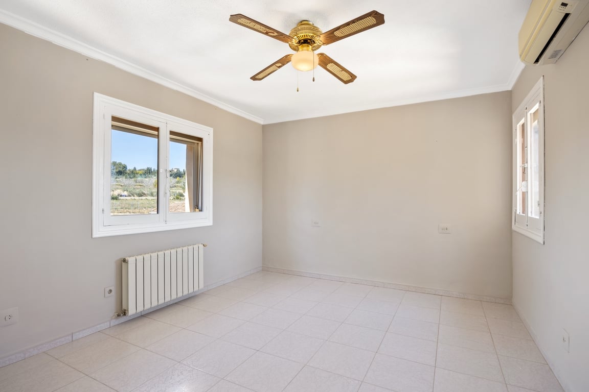 Helles Schlafzimmer mit Fliesenboden, klassischem Deckenventilator, Klimaanlage und Heizkörper. Die Fenster bieten einen weiten Blick auf die umliegende Landschaft.
