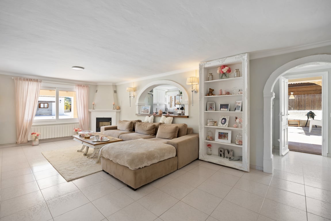 Helles Wohnzimmer mit Eckkamin, Modulsofa und Rundbogenöffnungen zur Küche und Terrasse. Ausgestattet mit einem dekorativen Regal und hellem Fliesenboden.