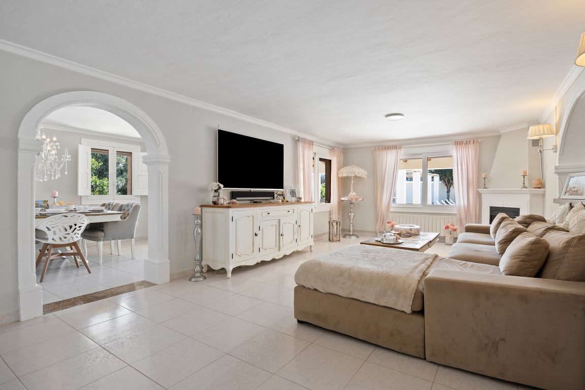 Helles Wohnzimmer mit Keramikböden, Ecksofa und Kamin. Ein dekorativer Rundbogen führt zum Essbereich und unterstreicht die offene Raumaufteilung und das Tageslicht.