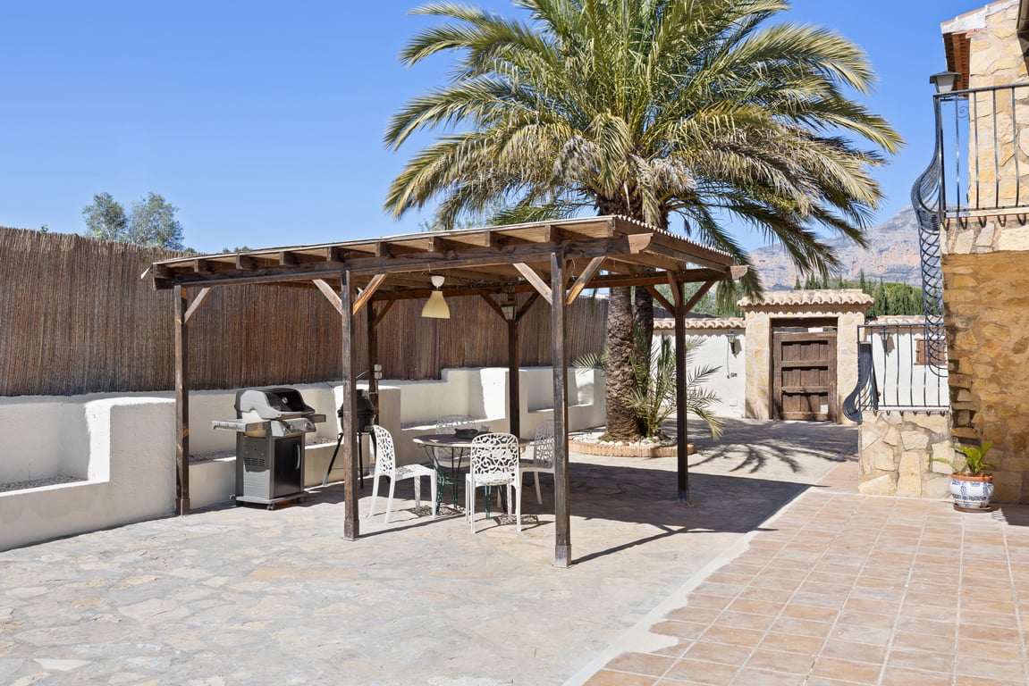 Holzpergola mit Außen-Essbereich und Grill auf einer Natursteinterrasse, umgeben von einer großen Palme und einer Steinmauer mit Bergblick.