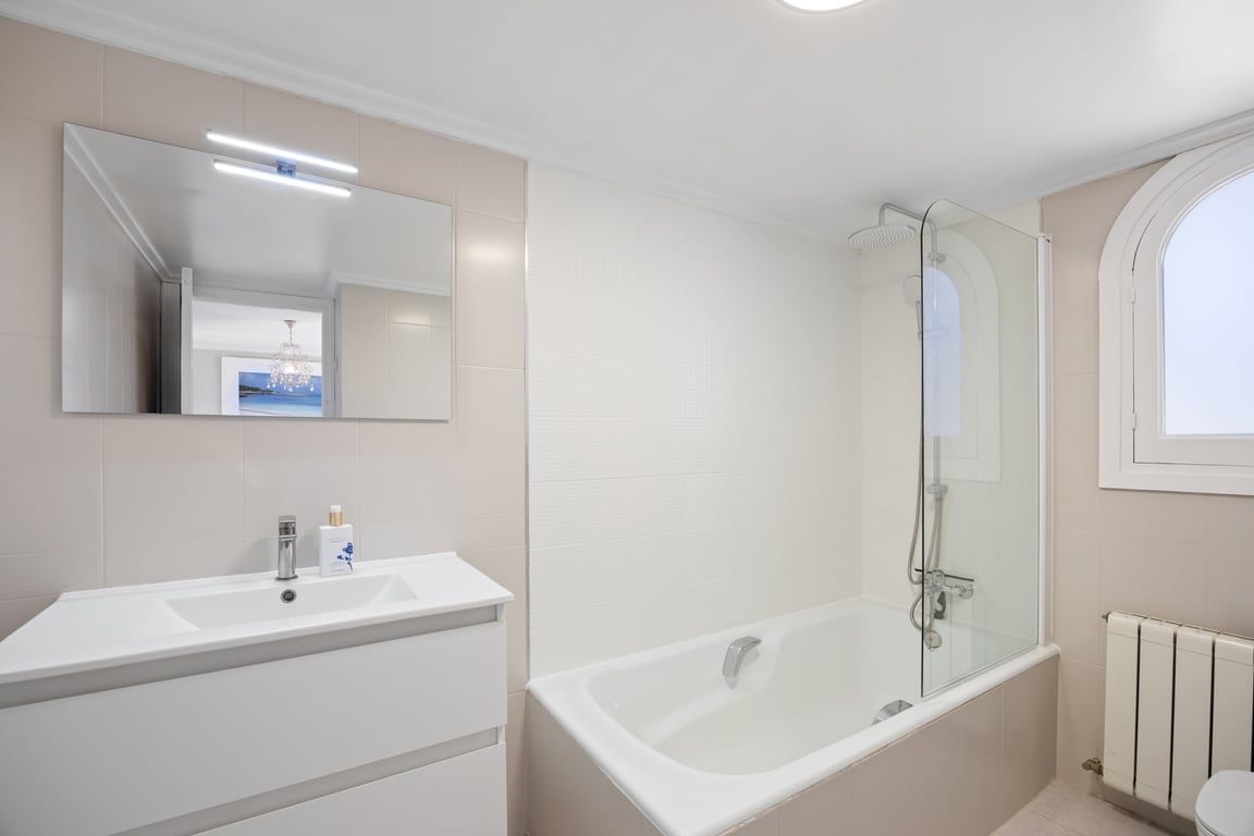 Modernes Badezimmer mit weißem Waschtisch, beleuchtetem Spiegel und Badewanne mit Glasduschwand. Ein Rundbogenfenster sorgt für natürliches Licht neben neutralen Fliesen.