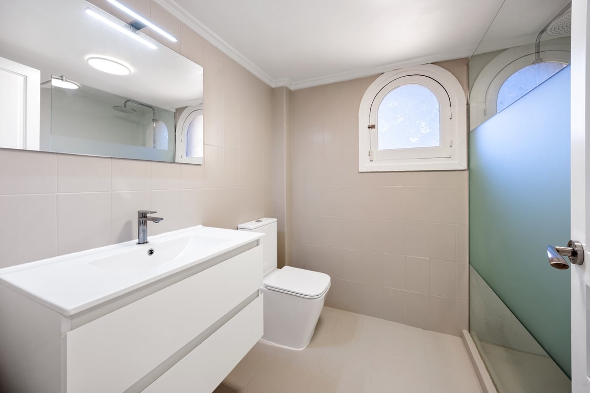 Modernes Badezimmer mit weißem Waschtisch, Dusche mit Glaswand und einem charakteristischen Rundbogenfenster. Neutrale Fliesen und helle LED-Beleuchtung sorgen für Komfort.