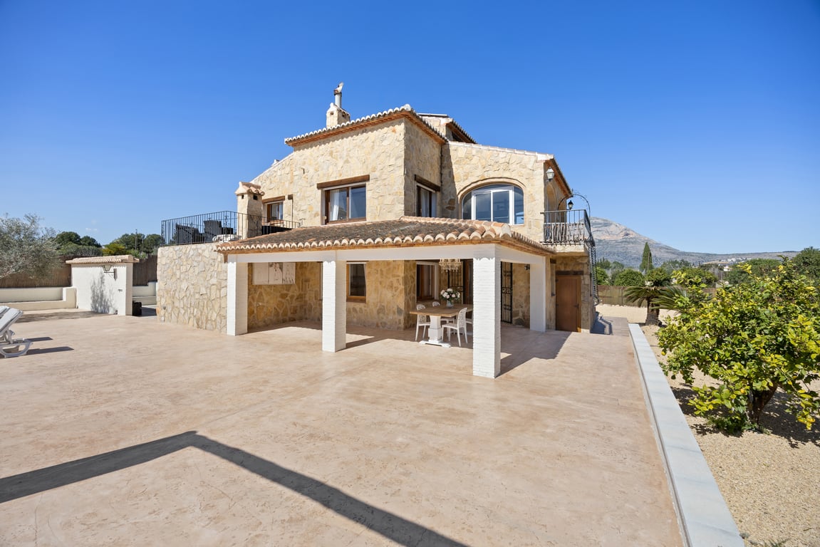 Natursteinfassade mit überdachter Naya, weißen Säulen und weitläufiger gepflasterter Terrasse mit Blick auf den Berg Montgó in Jávea.